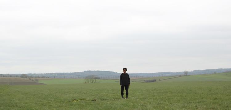 Photo d'une personne noire, habillé en noir et qui se tient debout dans une prairie